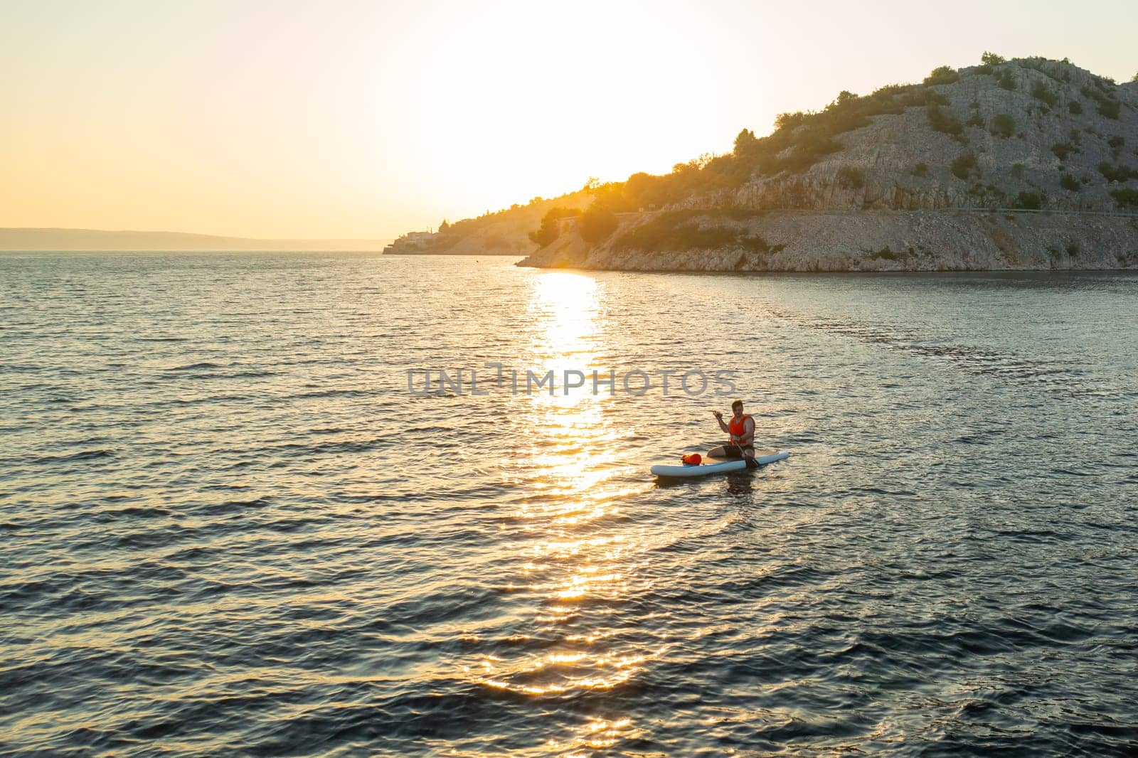Man in an orange vest sits on his paddleboard, moving across the serene sea at sunset