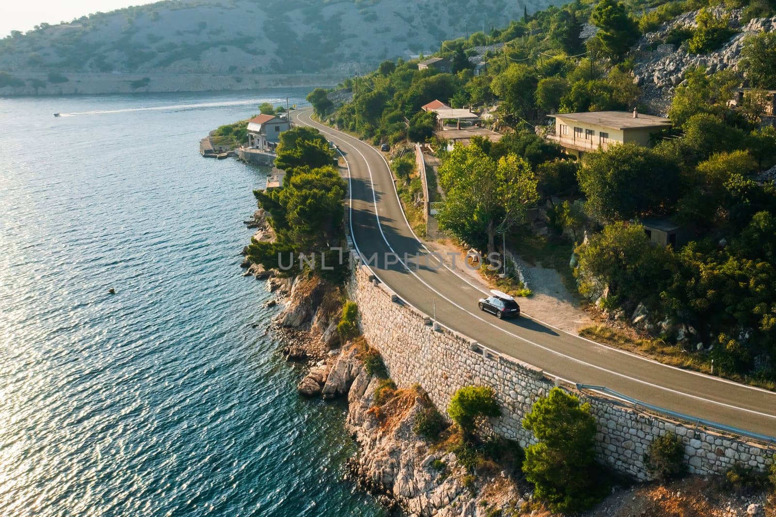 Car with paddleboard on the roof drives along a narrow coastal road under a clear blue sky.