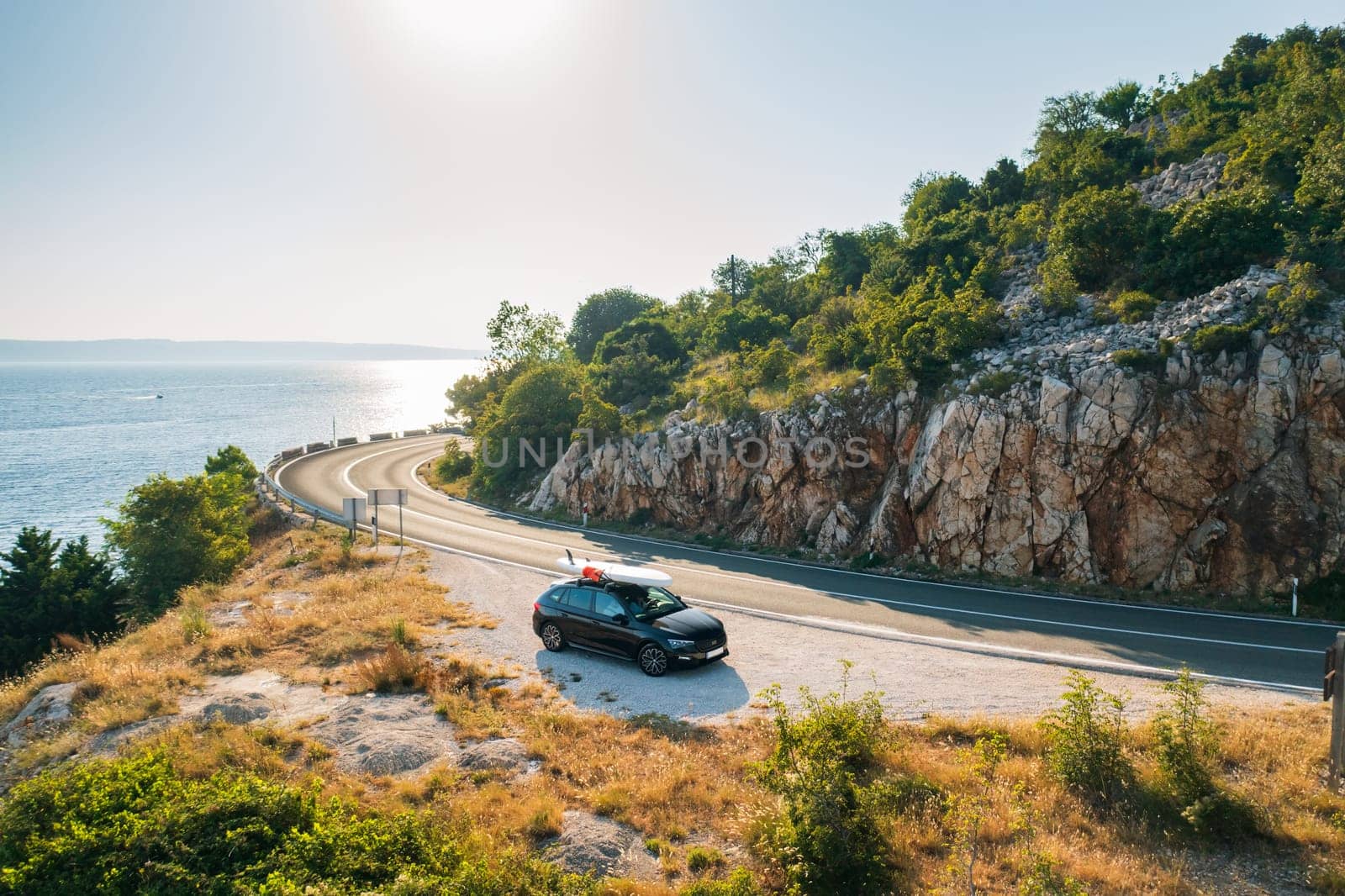 Car with a SUP lashed onto its roof, parked on the side of a quiet country road, with the shimmering sea visible through the trees