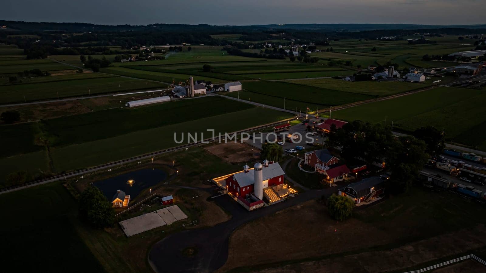 Ronks, Pennsylvania, USA, July 19 2024 - Tranquil Farm Scene At Dusk Featuring A Large Red Barn With A White Silo, An Illuminated Stone Courtyard, A Nearby Brick House, Expansive Green Fields, A Reflective Pond, And A Scenic View Of The Surrounding Countryside Under A Softly Lit Evening Sky.