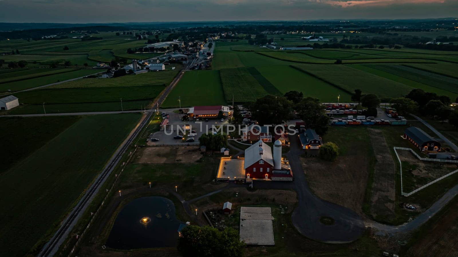 Ronks, Pennsylvania, USA, July 19 2024 - Tranquil Farm Scene At Dusk Featuring A Large Red Barn With A White Silo, An Illuminated Stone Courtyard, A Nearby Brick House, Expansive Green Fields, A Reflective Pond, And A Scenic View Of The Surrounding Countryside Under A Softly Lit Evening Sky.