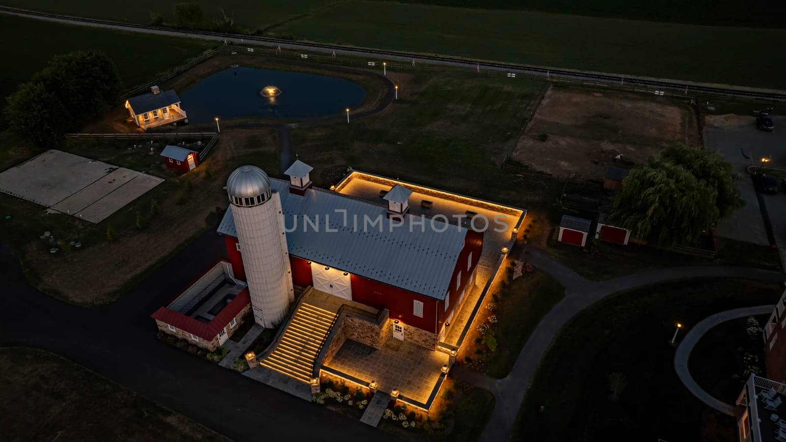 Ronks, Pennsylvania, USA, July 19 2024 - Tranquil Farm Scene At Dusk Featuring A Large Red Barn With A White Silo, An Illuminated Stone Courtyard, A Nearby Cozy Stone House Reflecting In A Pond, Expansive Green Fields, And A Serene Evening Sky