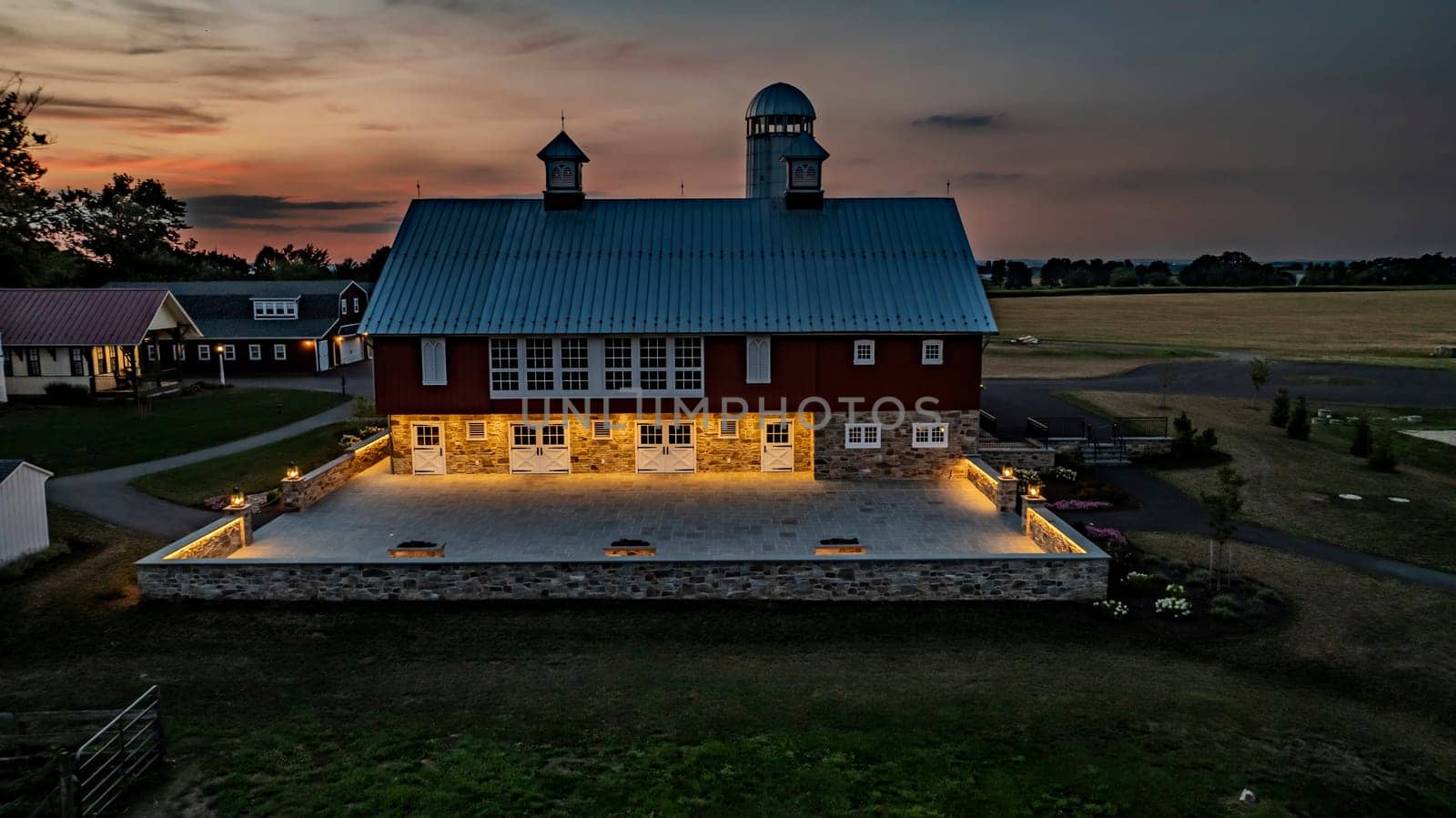 Ronks, Pennsylvania, USA, July 19 2024 - Peaceful Farm Scene At Dusk Featuring A Large Red Barn With A White Silo, An Illuminated Stone Courtyard, Adjacent Buildings, And Expansive Green Fields Under A Beautifully Lit Sunset Sky.