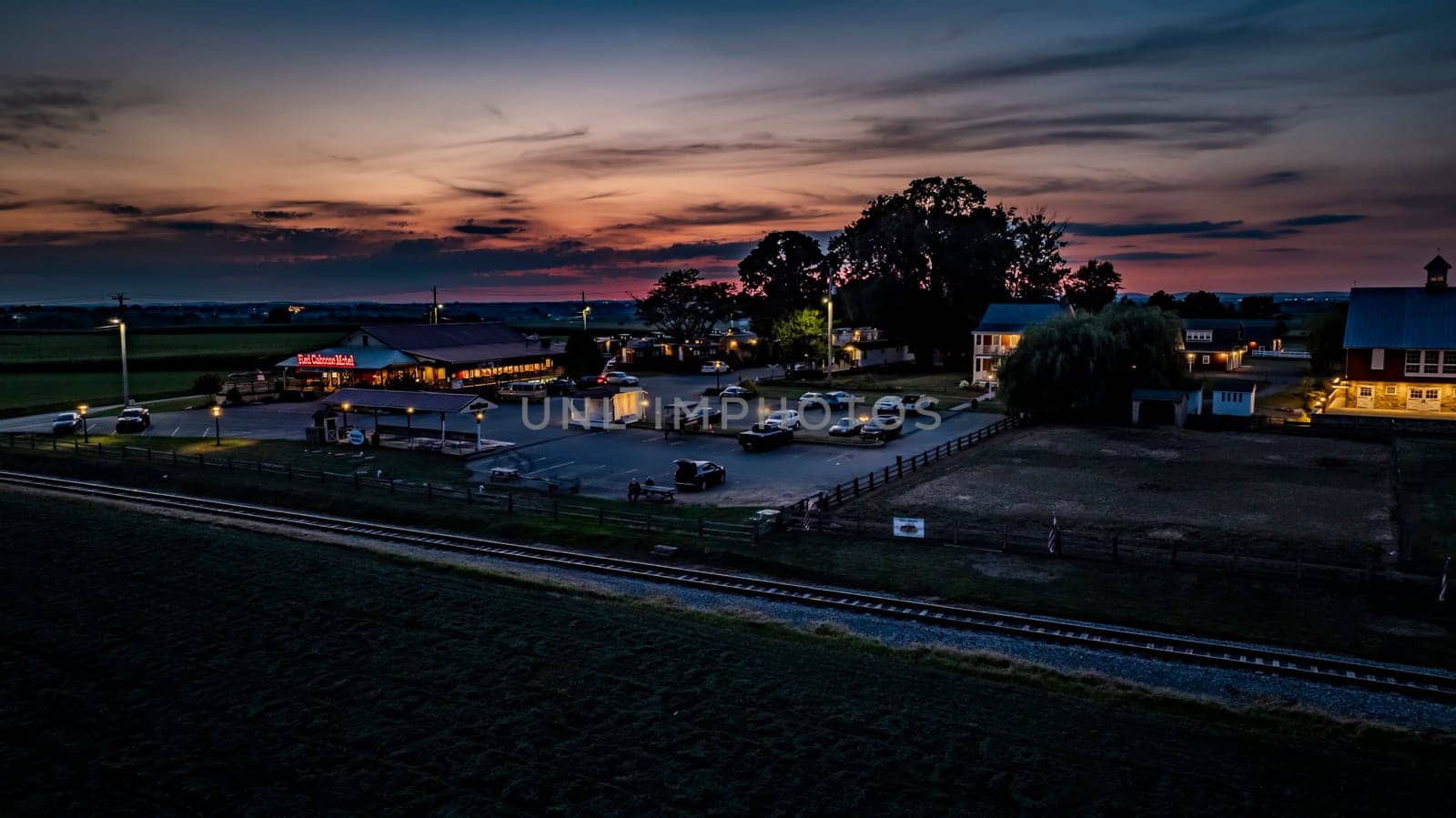 Ronks, Pennsylvania, USA, July 19 2024 - Tranquil Evening Scene Featuring A Farm With A Red Barn, Cozy Buildings, A Parking Lot With Cars, Illuminated By Soft Lighting, A Nearby Railroad Track, And A Vibrant Sunset Sky Creating A Serene Atmosphere.