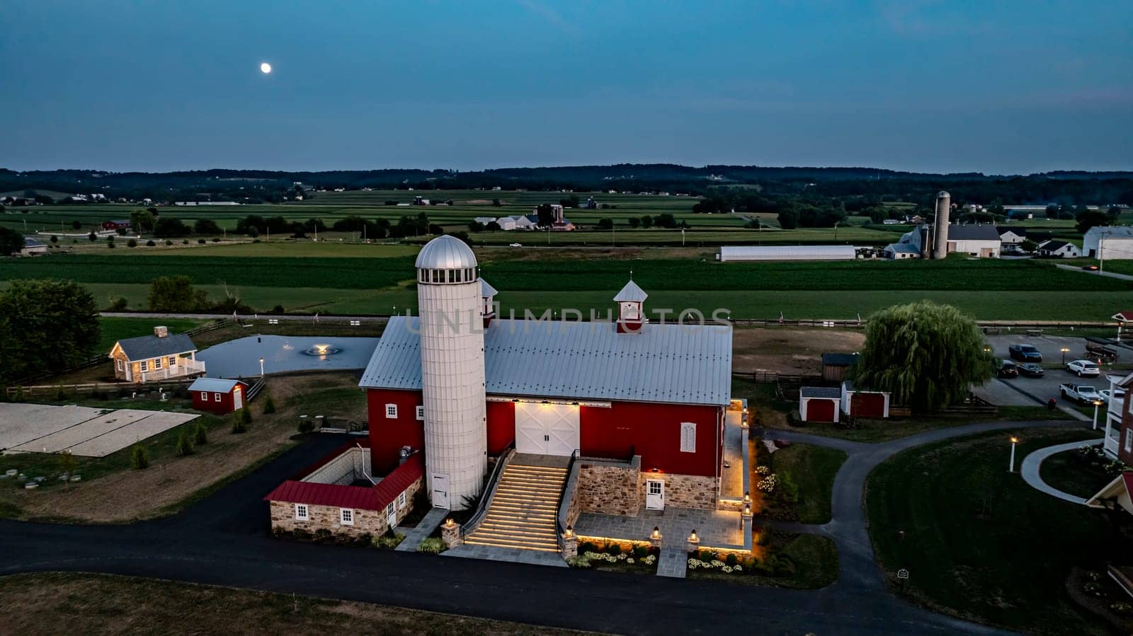 Ronks, Pennsylvania, USA, July 19 2024 - Serene Farm Scene Dusk Featuring Large Red Barn With Silo, Stone Courtyard Illuminated By Soft Lighting, Charming Stone House Reflecting In Pond, Expansive Green Fields, Rising Full Moon.