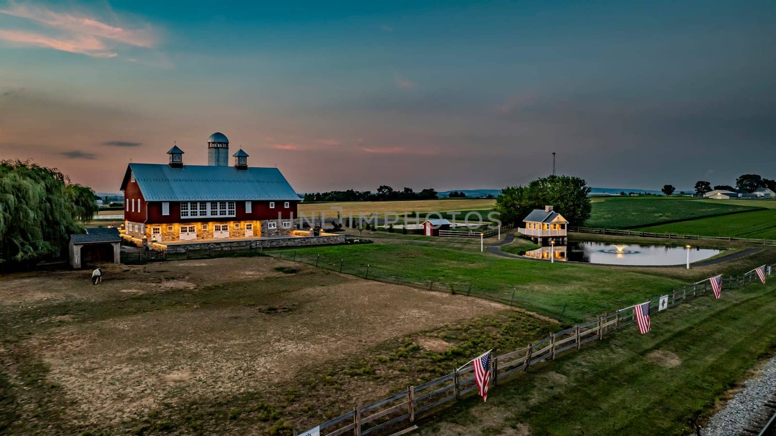 Ronks, Pennsylvania, USA, July 19 2024 - Peaceful Farm Scene At Dusk Featuring A Large Red Barn With A White Silo, A Charming Stone House Reflecting In A Pond, Expansive Green Fields, And A Softly Lit Evening Sky