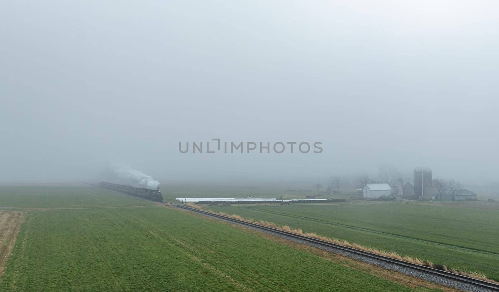 A steam train makes its way through a foggy landscape, surrounded by green fields and distant farm buildings.