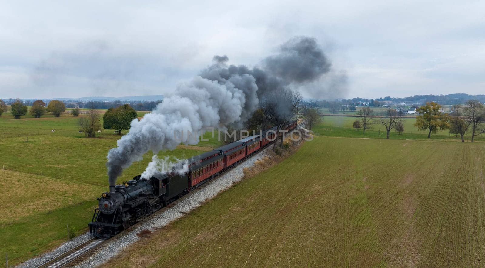 A classic steam train chugs along the tracks, billowing smoke as it journeys through green fields under an overcast sky.