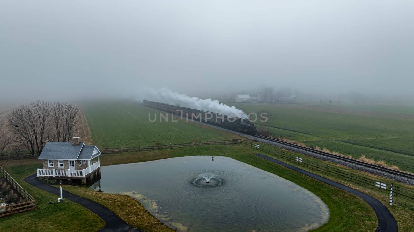 A steam train navigates through fog-covered fields beside a quaint house and a pond, creating a serene atmosphere.