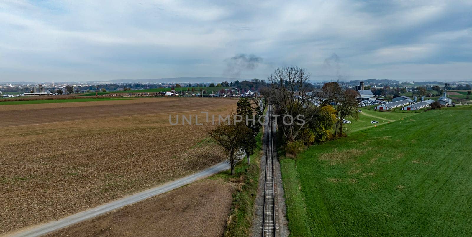A vast expanse of farmland stretches alongside railway tracks, leading toward a town beneath a cloudy sky.