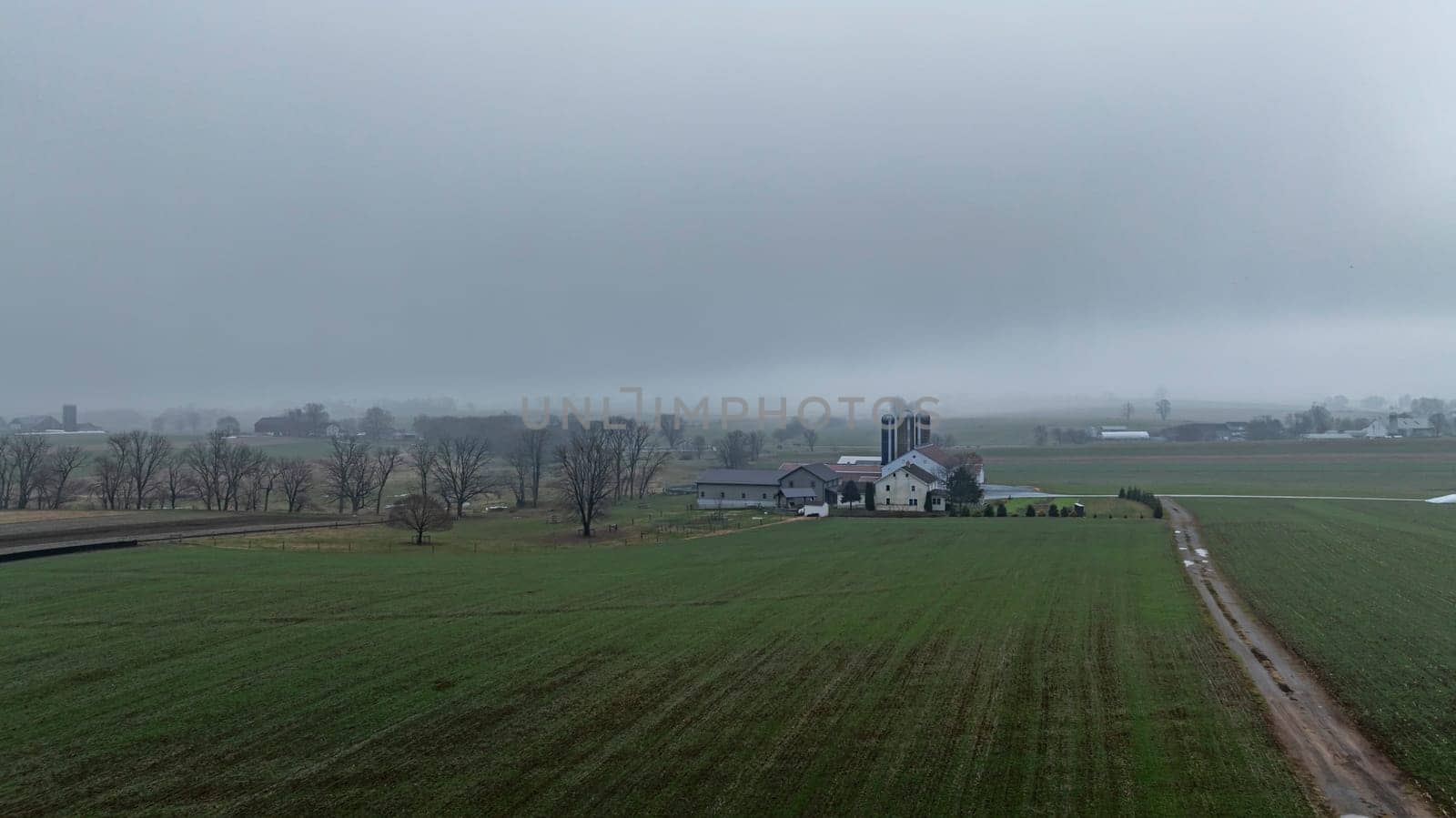 A rural farmstead is enveloped in fog, surrounded by expansive green fields and distant structures on a calm morning.