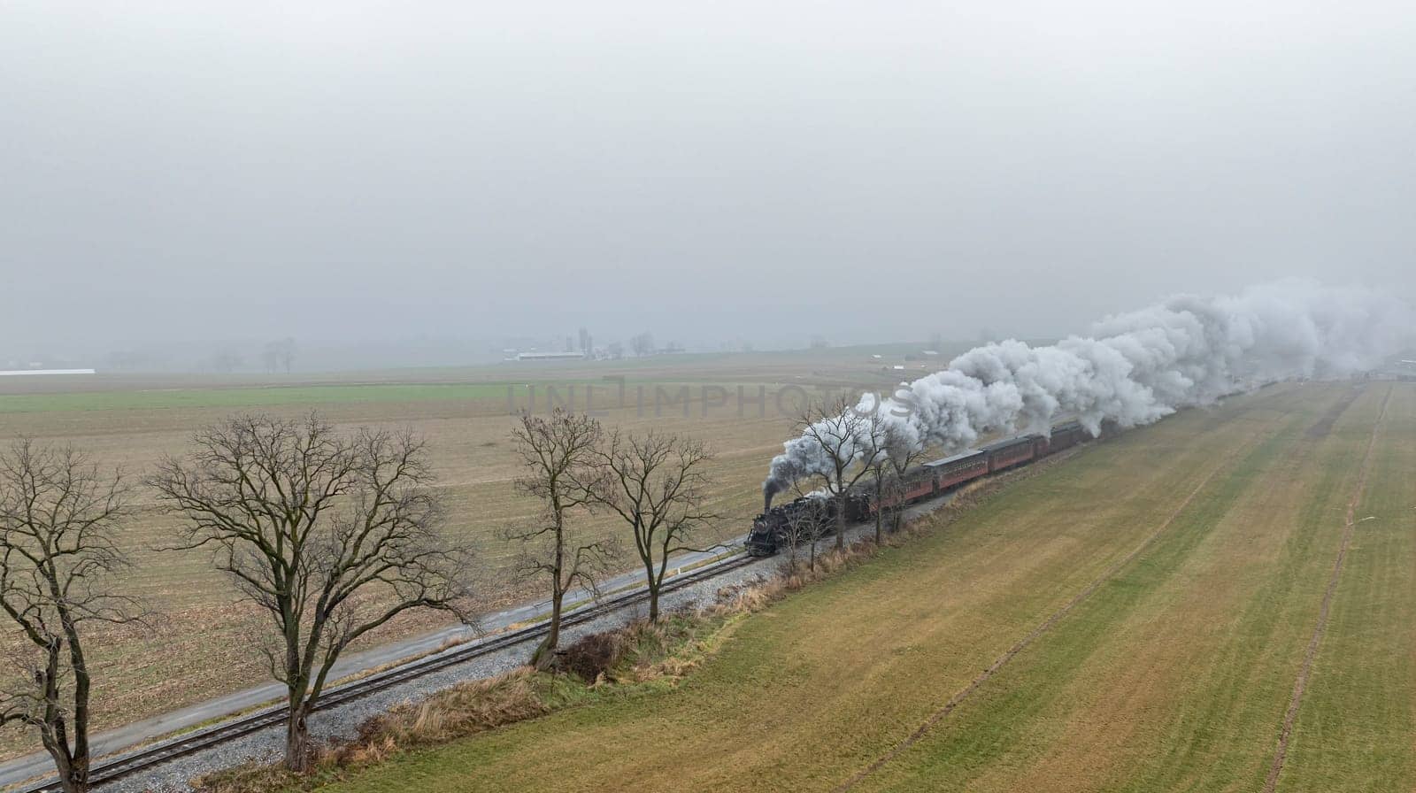 A steam train puffs clouds of smoke while traveling alongside fog-covered fields under an overcast sky.