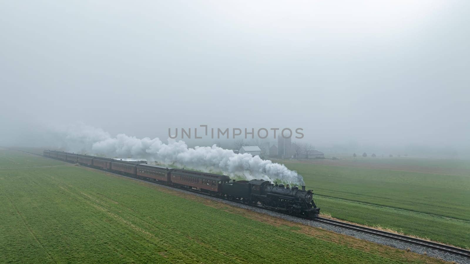 A steam locomotive pulls several carriages along a misty track, passing through lush green fields, with farm buildings faintly visible in the background.