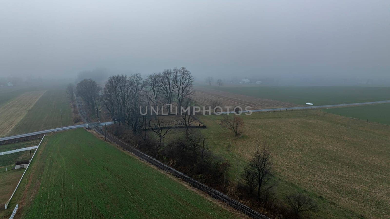 A misty morning covers a rural area, highlighting scattered trees and expansive fields, with a road partially visible.