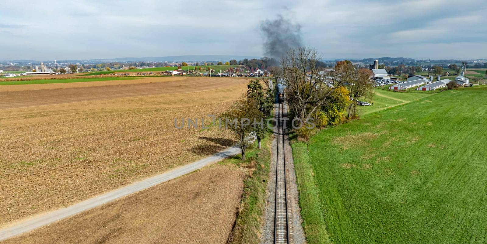 A train moves along tracks surrounded by fields and trees, with a rural town visible in the background under gray skies.