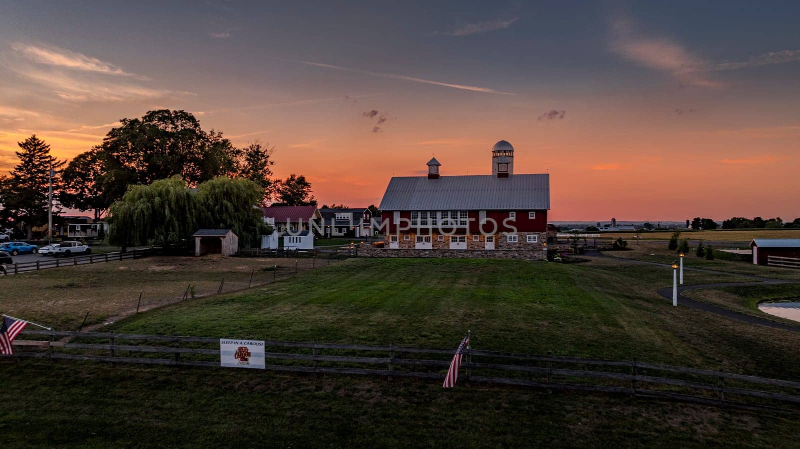 Ronks, Pennsylvania, USA, July 19 2024 - Large Red Barn With Silver Roof And Twin Cupolas At Sunset, Surrounded By Open Fields, A Weeping Willow, Fencing, And Other Farm Structures Under A Pink And Orange Sky.