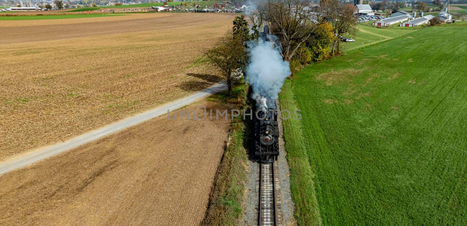 A vintage steam train chugs along grassy fields and farmland, surrounded by vibrant autumn colors.