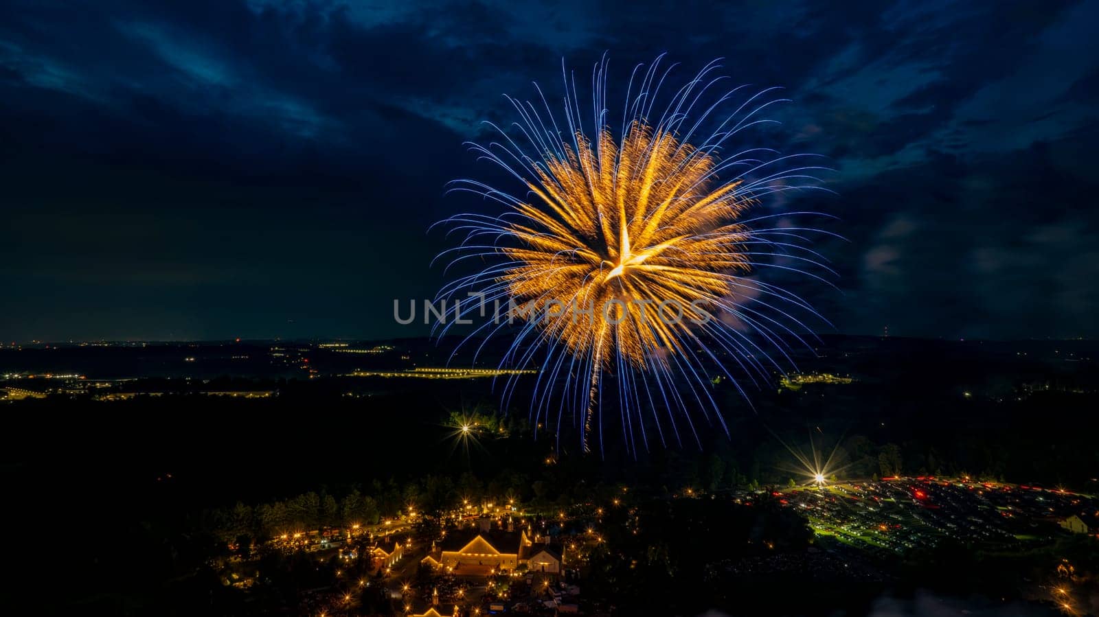 Golden And Blue Fireworks Burst Over A Well-Lit Estate, Surrounded By Trees And Fields, With Distant City Lights Adding Depth To The Festive Nighttime Scene.