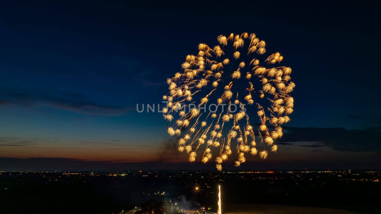 Golden Fireworks Exploding In The Night Sky, Forming A Large, Brilliant Burst Over The Distant City Lights And A Gathered Crowd, With A Twilight Horizon Adding To The Festive Scene.