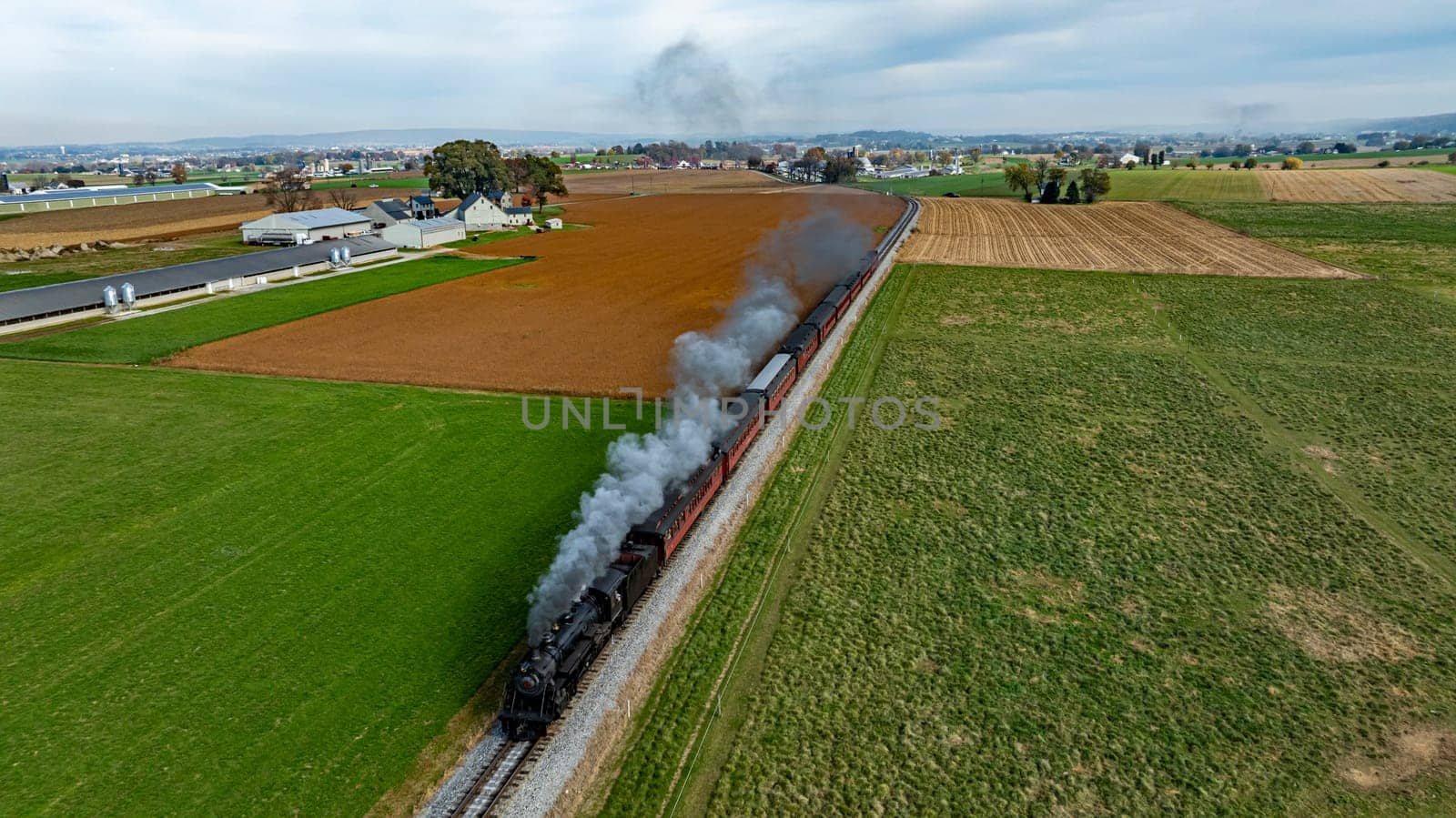 A steam train moves along the tracks surrounded by green fields and farmland under a cloudy sky.