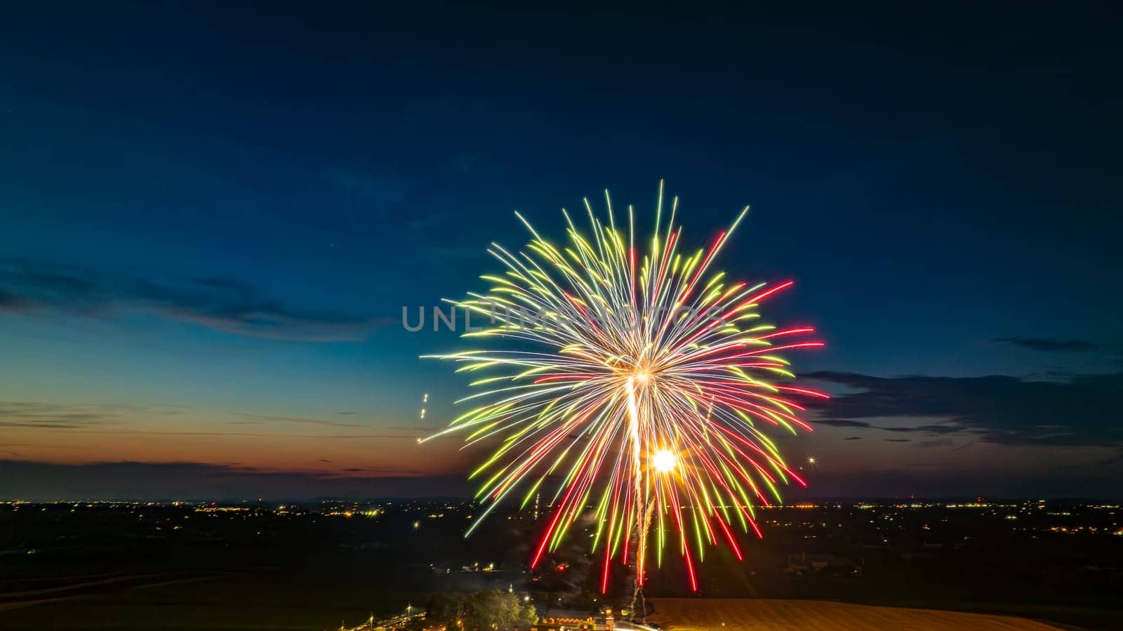 Colorful Fireworks Exploding In The Night Sky, Casting Bright Red, Green, And Yellow Bursts Over The Distant Cityscape And A Gathered Crowd, With The Twilight Horizon Adding To The Festive Atmosphere.