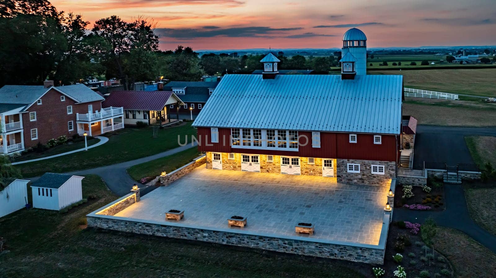 Ronks, Pennsylvania, USA, July 19 2024 - Tranquil Farm Scene At Dusk Featuring A Large Red Barn With A White Silo, An Illuminated Stone Courtyard, A Nearby Brick House, And Expansive Green Fields Under A Beautiful Sunset Sky, Creating A Serene And Picturesque Setting.