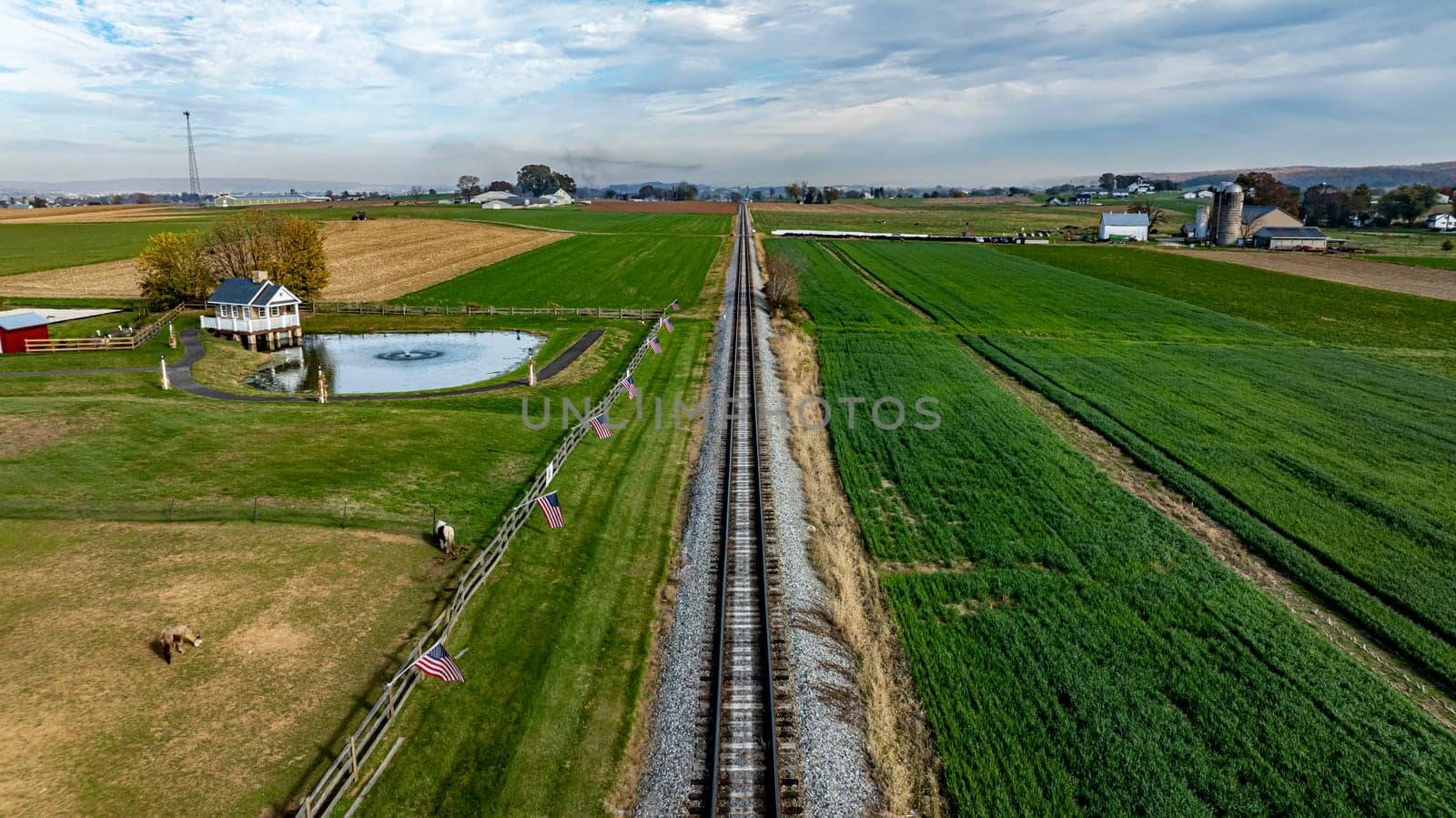 A serene rural landscape features railroad tracks leading through green fields and farms under a cloudy sky.