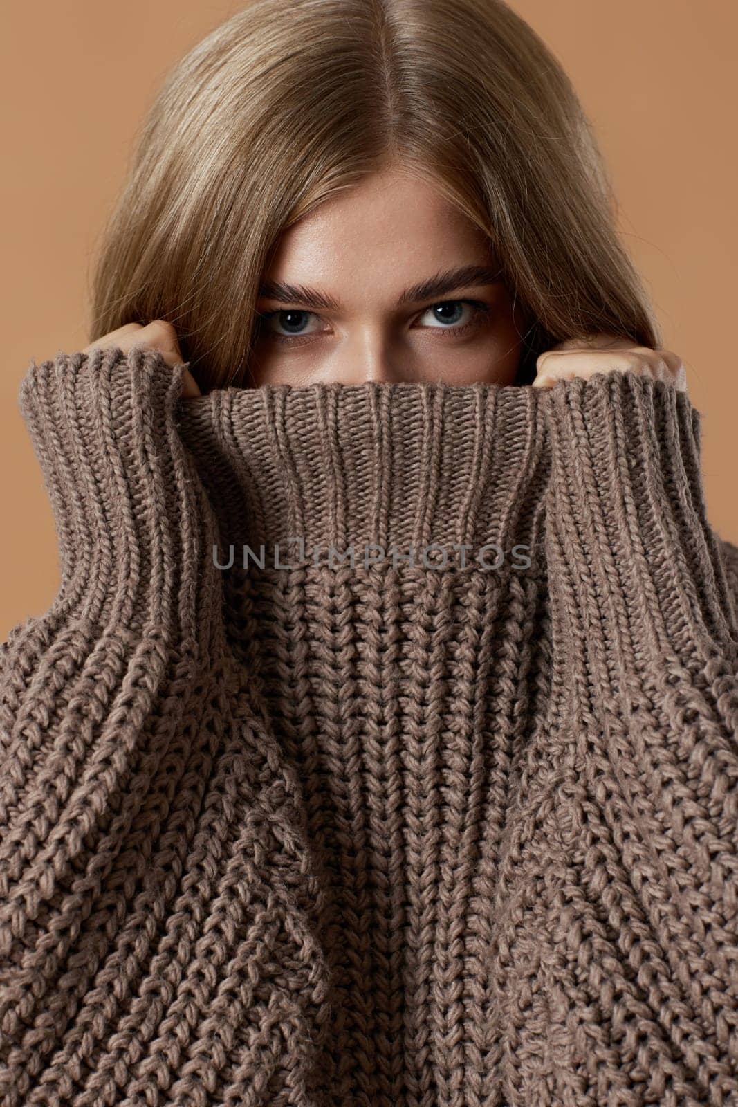 beautiful blonde woman covering face with turtleneck knitted sweater on beige background. autumn style