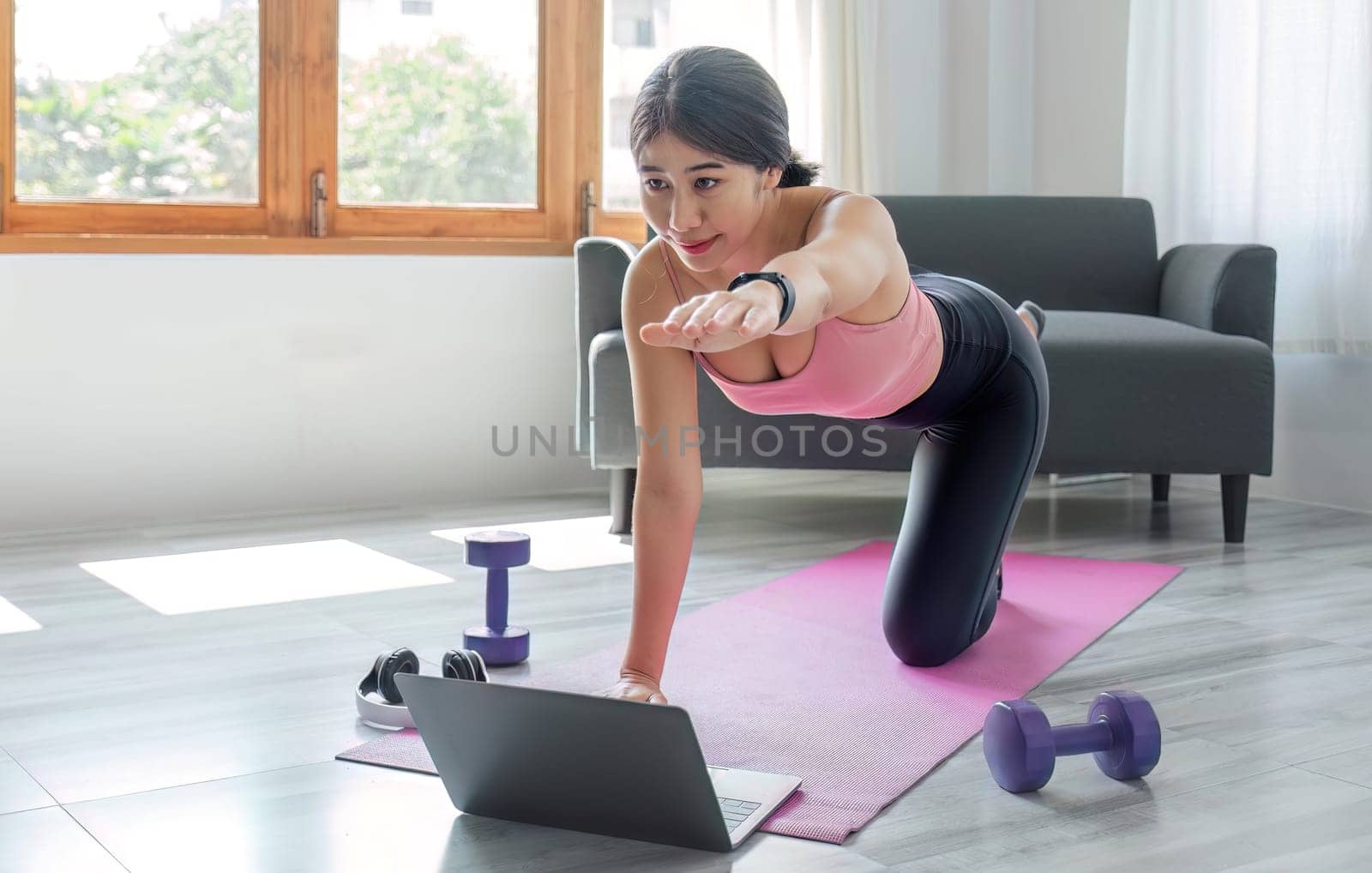 Young woman exercising in front of laptop Wear a sports bar outfit. Do yoga and lift dumbbells on the exercise mat. by wichayada