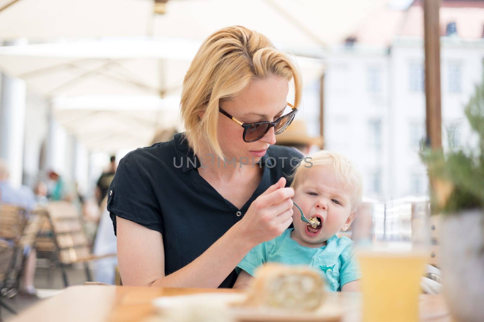 Young caucasian blonde mother spoon feeding her little infant baby boy child outdoors on restaurant or cafe terrace in summer.