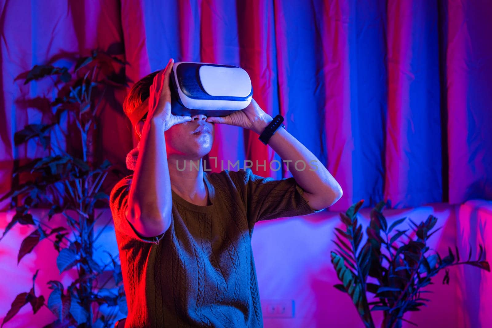 Asian woman wear the VR helmet excited emotional at home purple and blue background, female experiencing virtual reality goggles experiencing reality, metaverse and cyberspace concept