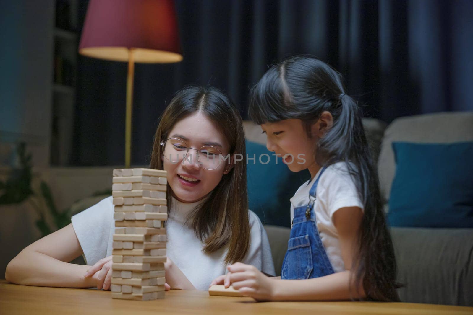 Asian young mother playing game in wood block with little daughter in home living room at night, Smiling woman help teach preschooler kid play build constructor tower of wooden blocks, family funny