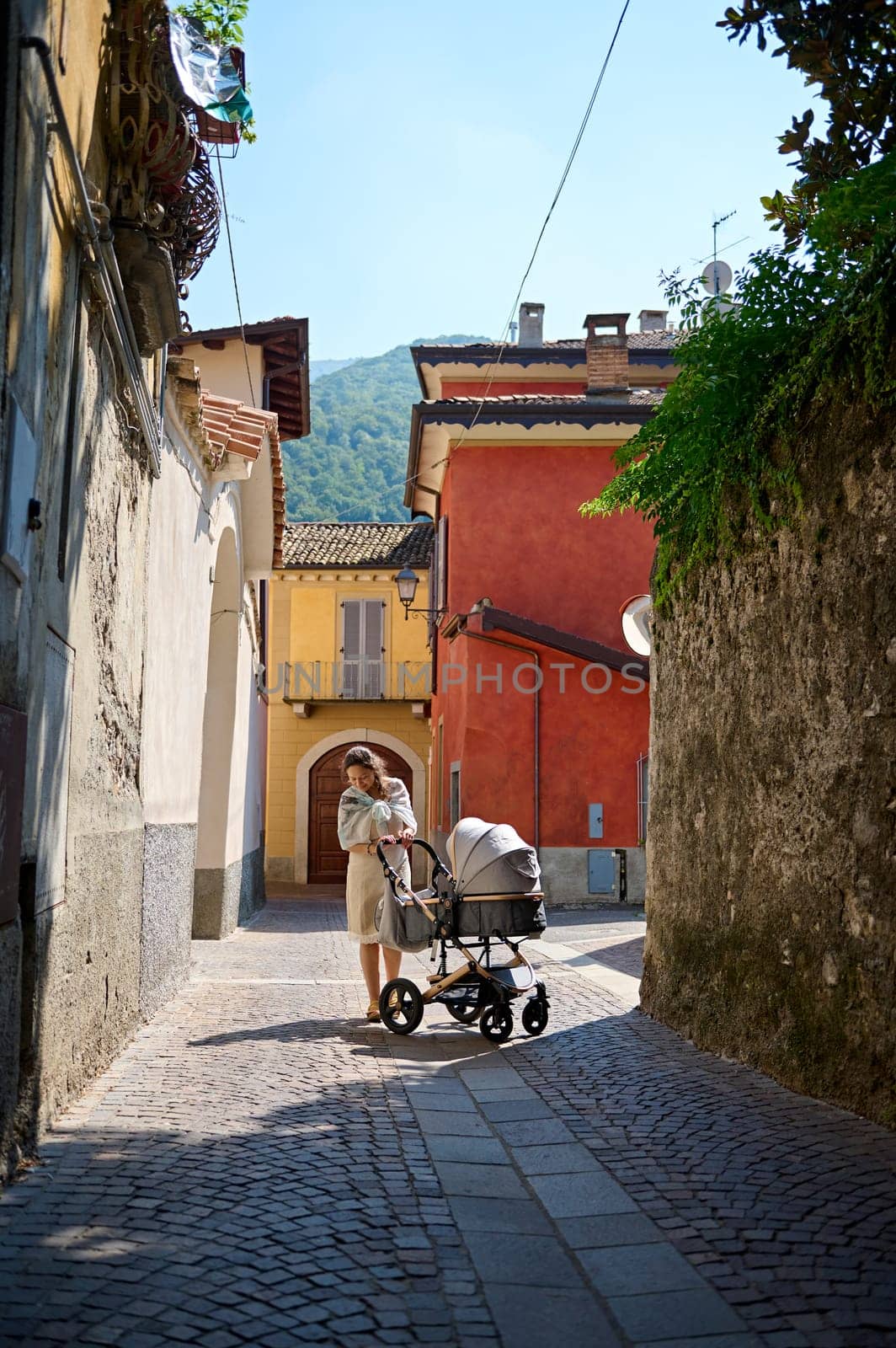 Full length portrait of a pretty woman in casual dress, standing with her baby in stroller in medieval cobblestone street. Inspired happy female tourist visiting Canzo. Lombardy, Italy. September 2023