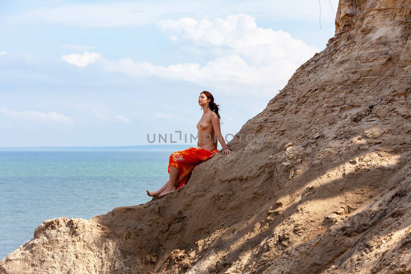 A young woman poses nude on a sand hill against a sky and seascape backdrop. Nature, beauty and nudity concept
