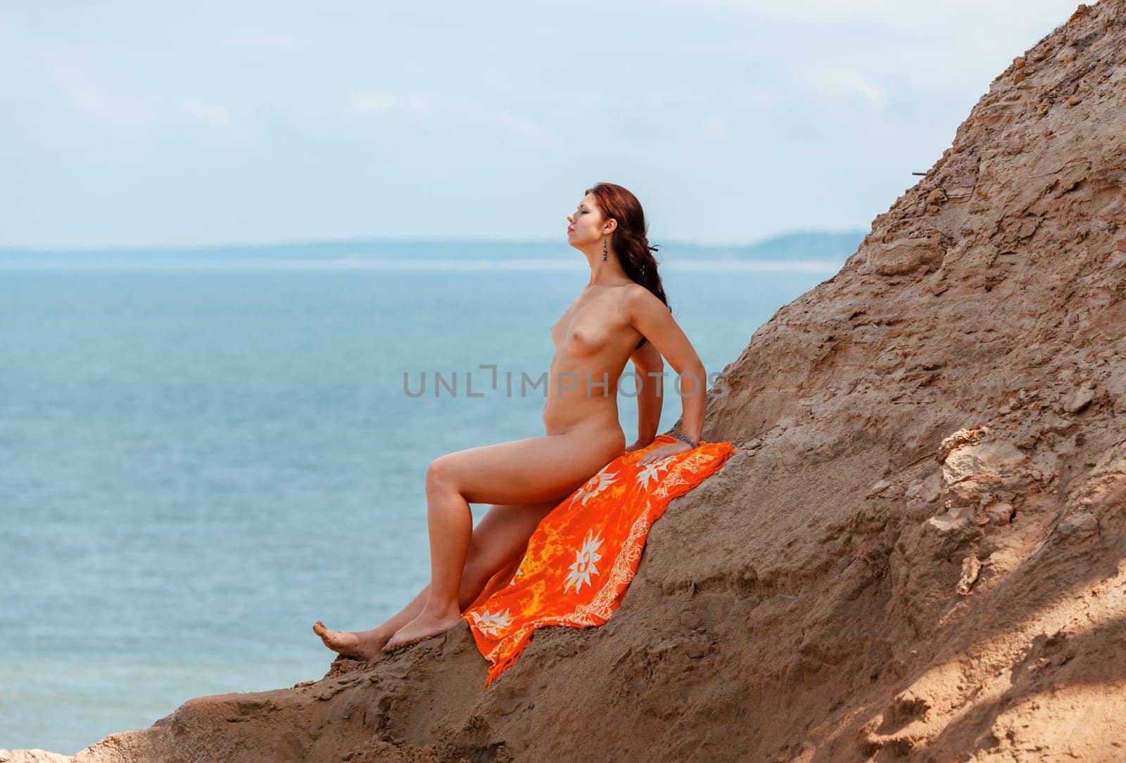 A young woman poses nude on a sand hill against a sky and seascape backdrop. Nature, beauty and nudity concept