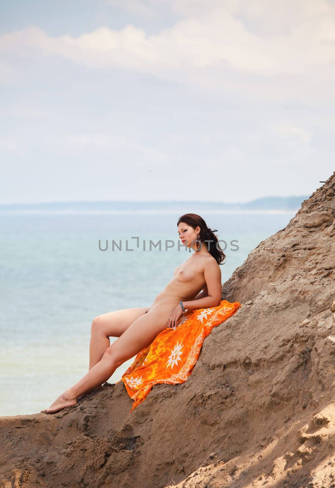 A young woman poses nude on a sand hill against a sky and seascape backdrop. Nature, beauty and nudity concept