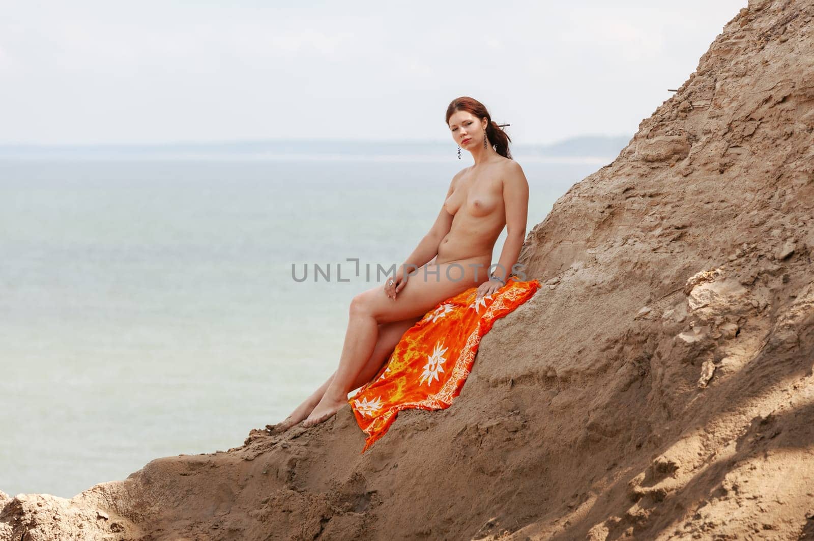 A young woman poses nude on a sand hill against a sky and seascape backdrop. Nature, beauty and nudity concept