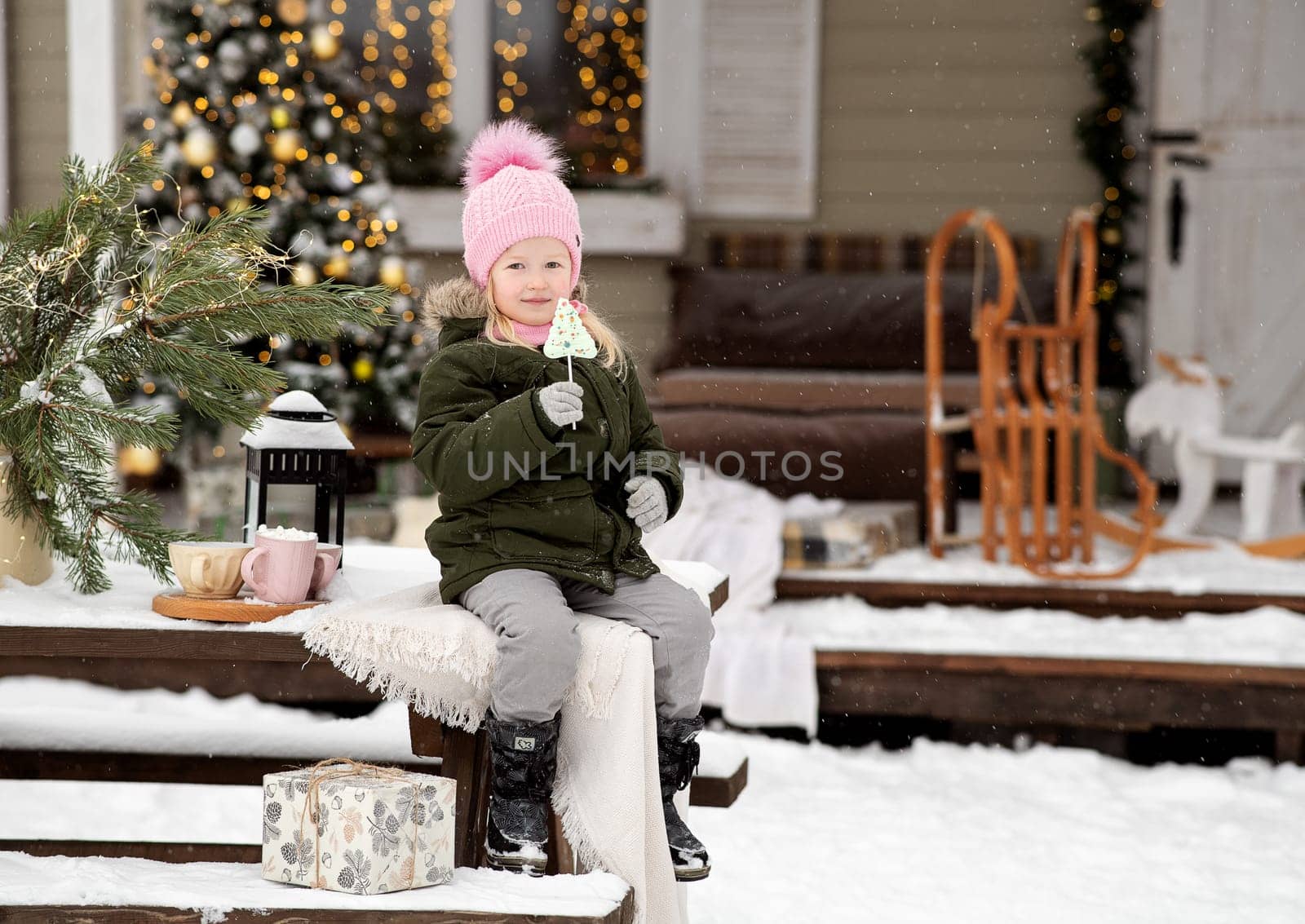A little girl in winter clothes is sitting on the porch of a house decorated for the Christmas holidays. Christmas holidays for a child