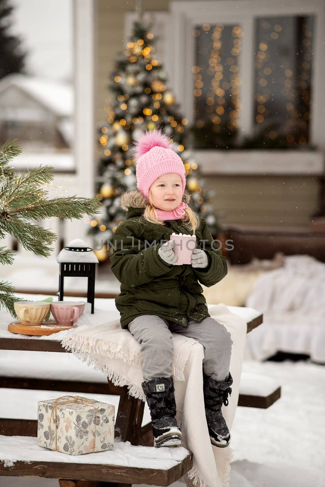 A little girl in winter clothes is sitting on the porch of a house decorated for the Christmas holidays. Christmas holidays for a child