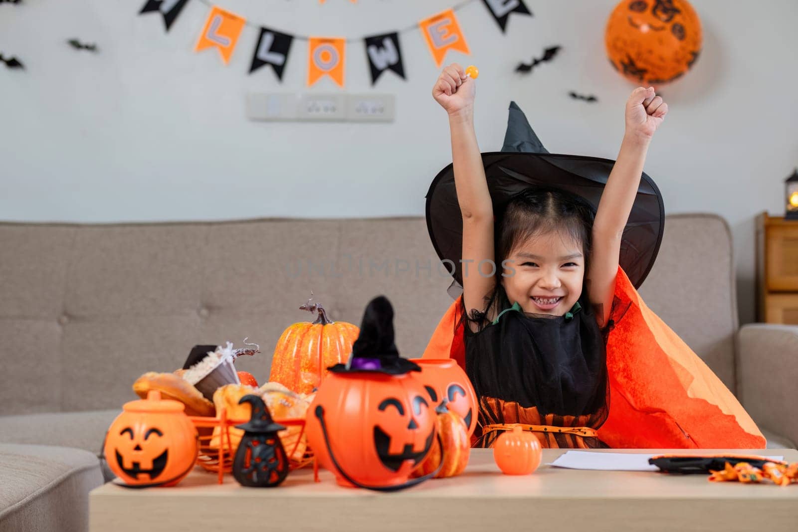 A cute young girl dressed in a witch costume enjoying Halloween festivities with pumpkins and spooky decorations in a cozy indoor setting.