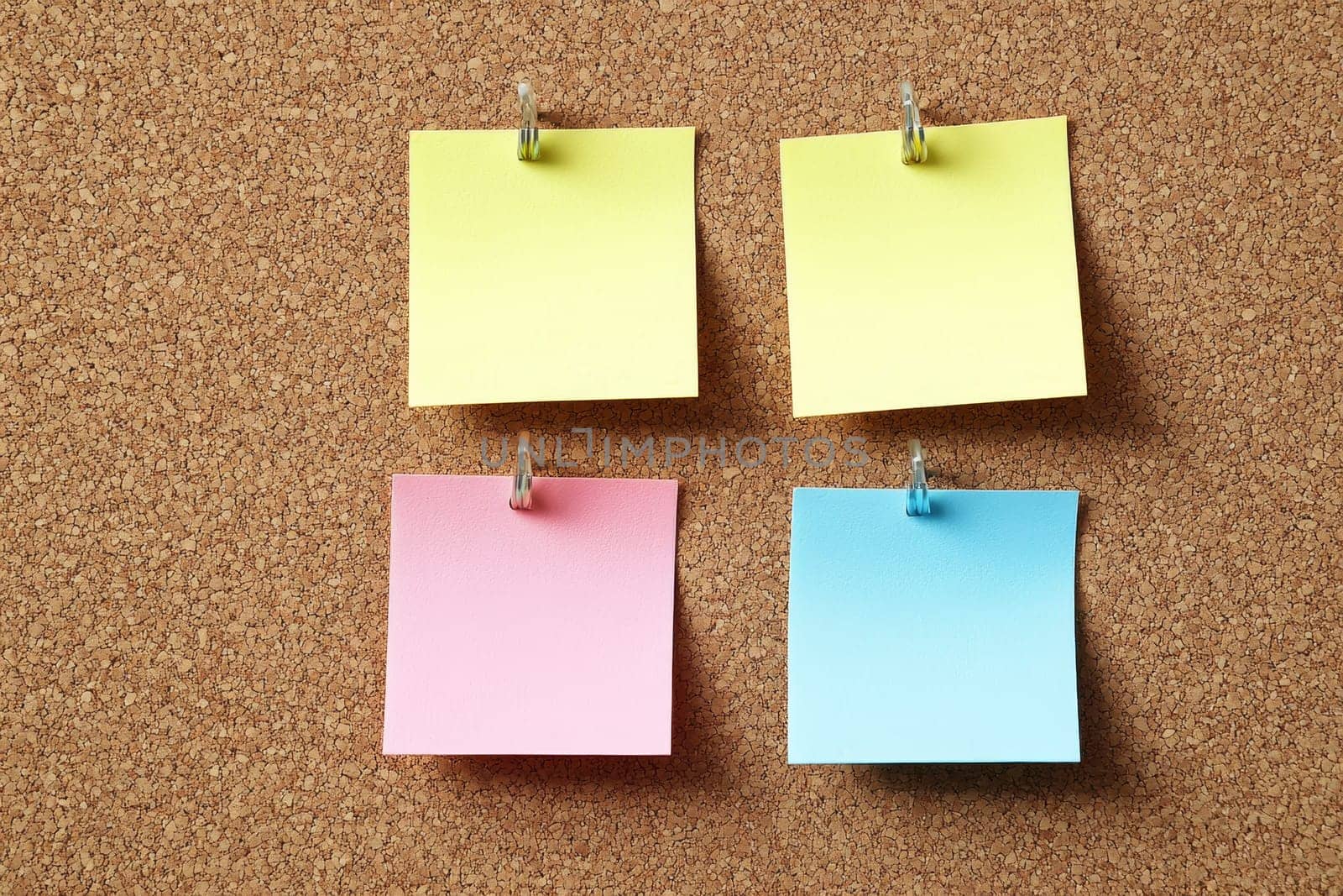 Four blank sticky notes in yellow, pink, and blue colors pinned in a grid pattern on cork board.