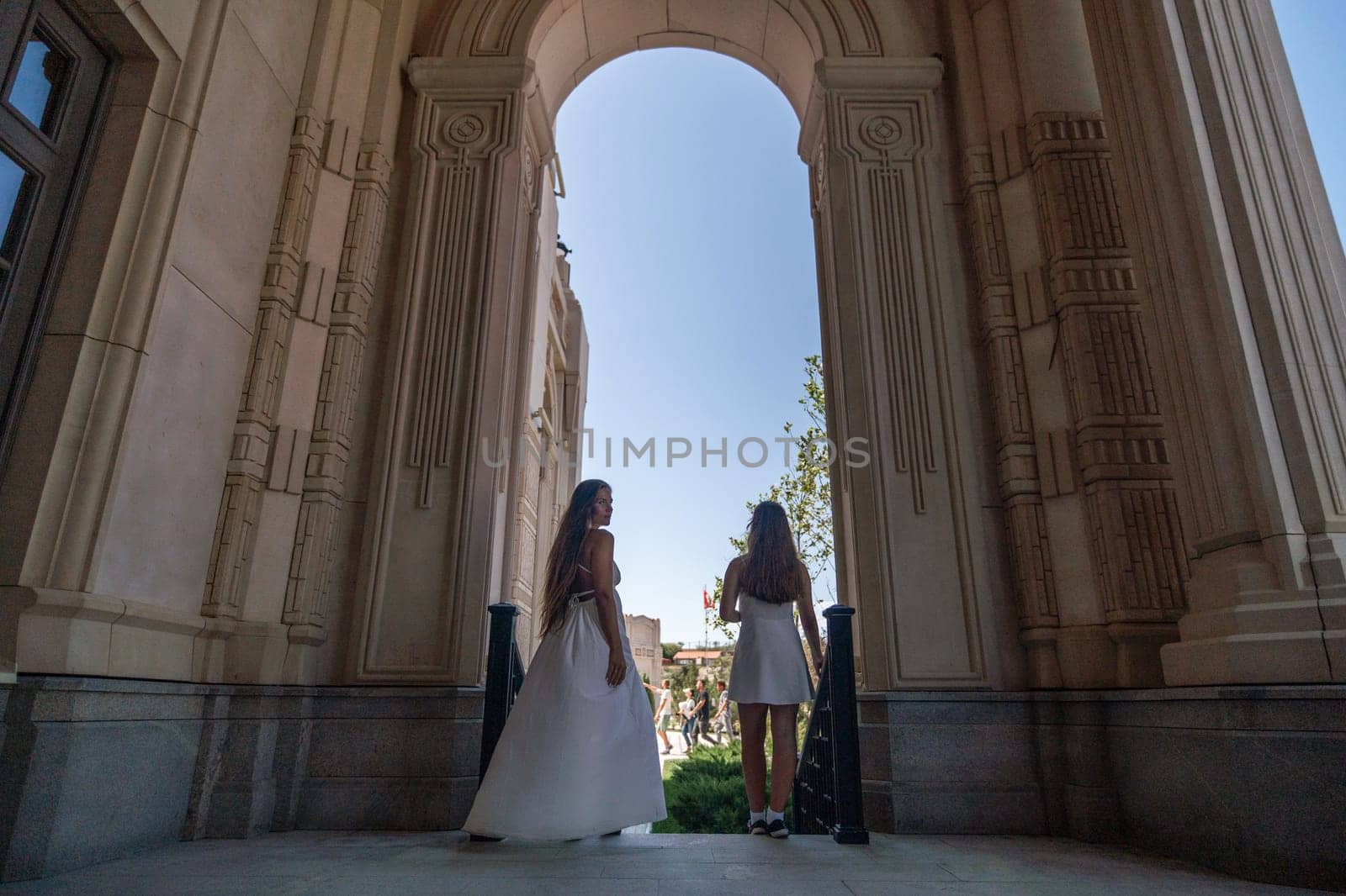 Two women in white dresses stand in an archway of a building, looking out at a clear blue sky.
