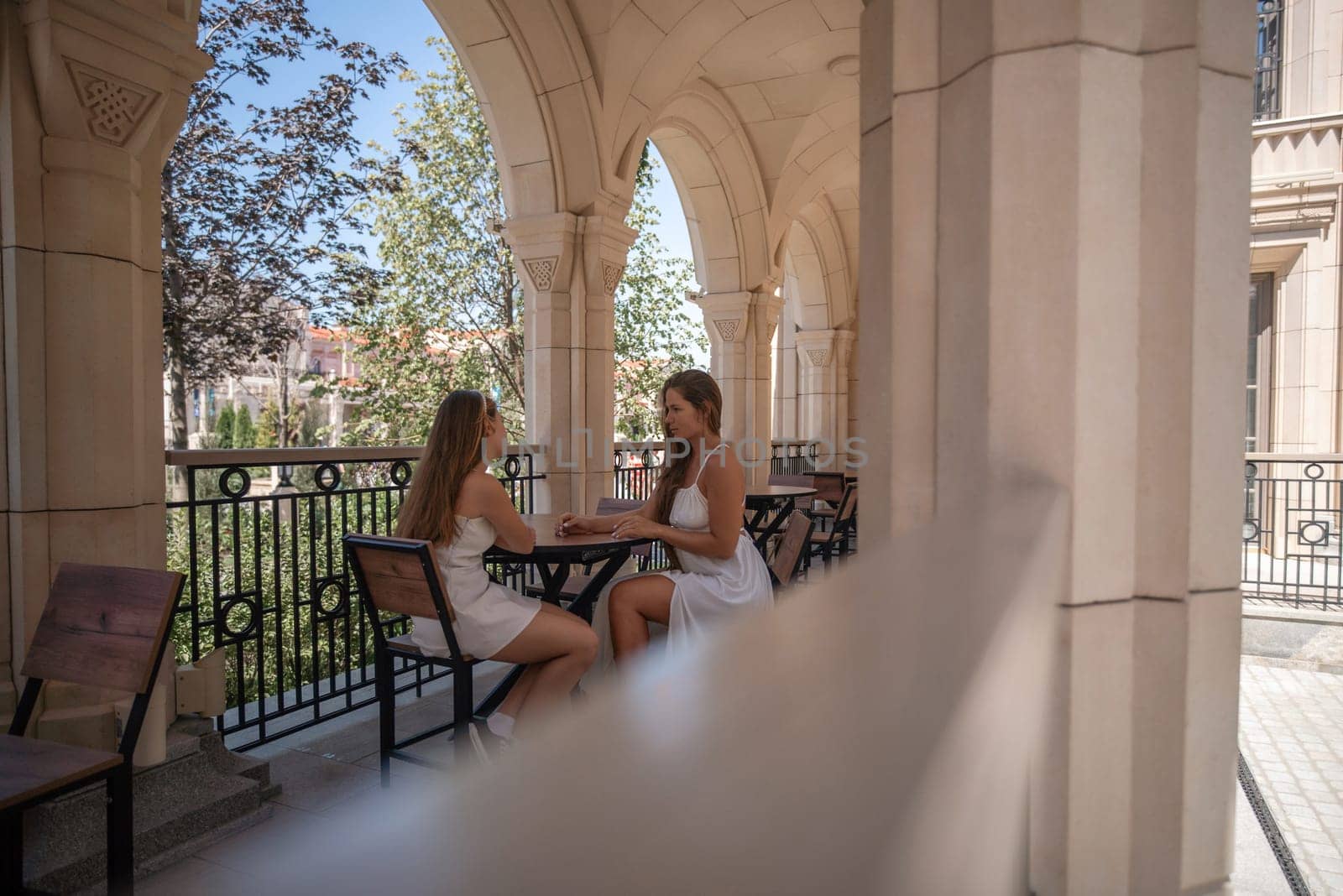 Two women in white dresses sit at a table on a balcony, chatting. The balcony has an ornate railing and is surrounded by arched columns.