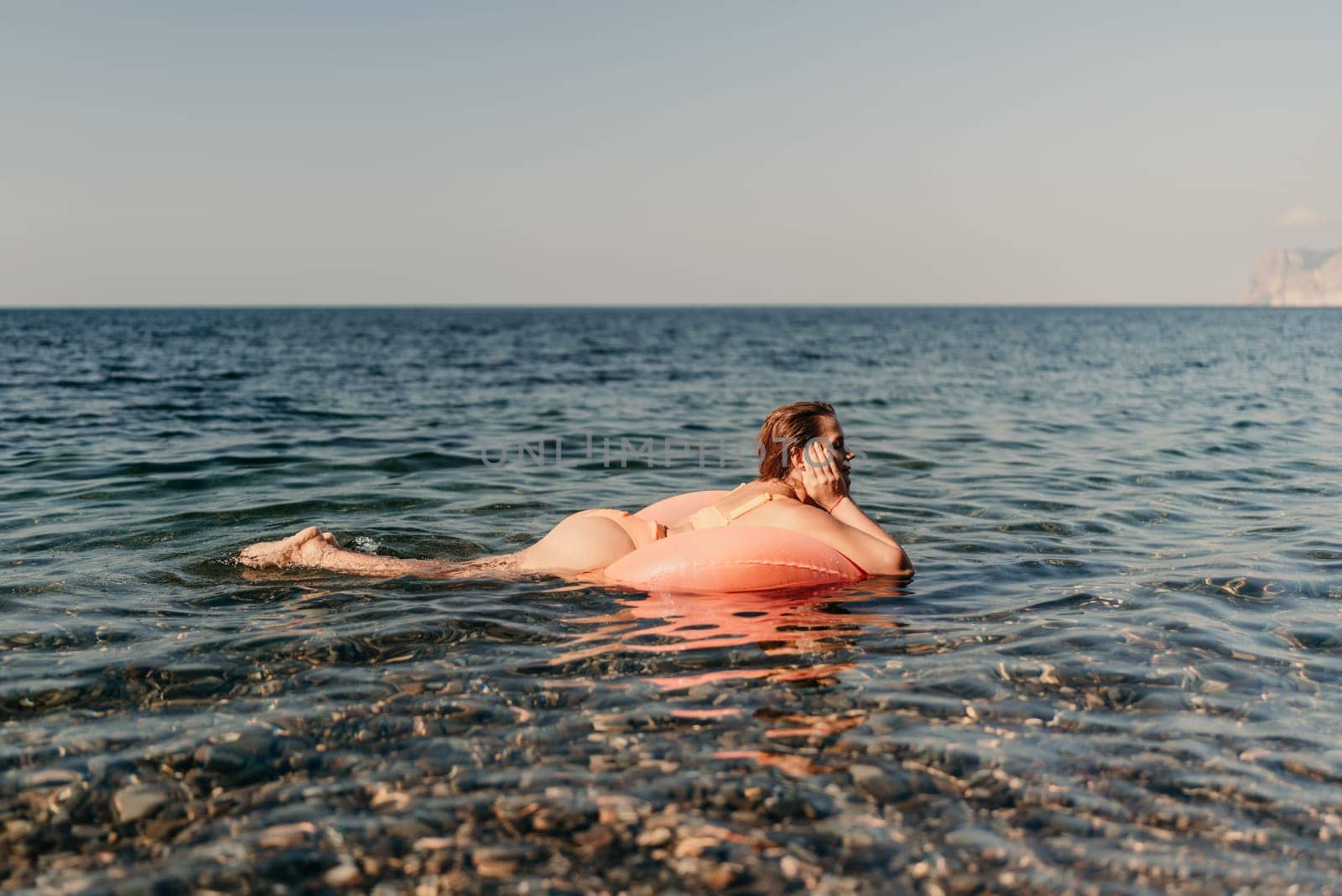 A woman is floating on a pink inflatable raft in the ocean. The water is calm and the sky is clear. The woman is enjoying her time in the water