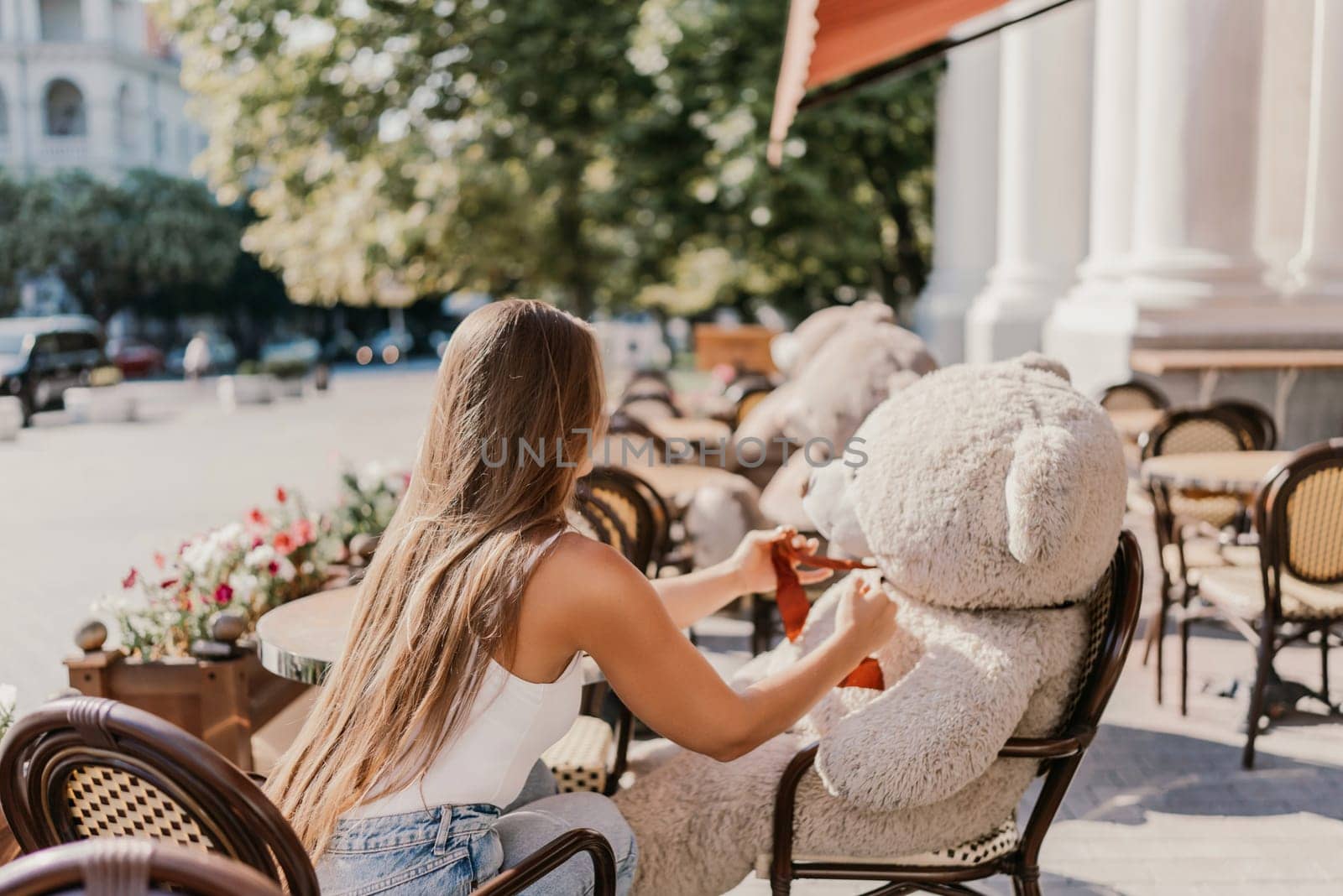 A woman sits cafe with a teddy bear next to her. The scene is set in a city with several chairs and tables around her. The woman is enjoying her time at the outdoor cafe