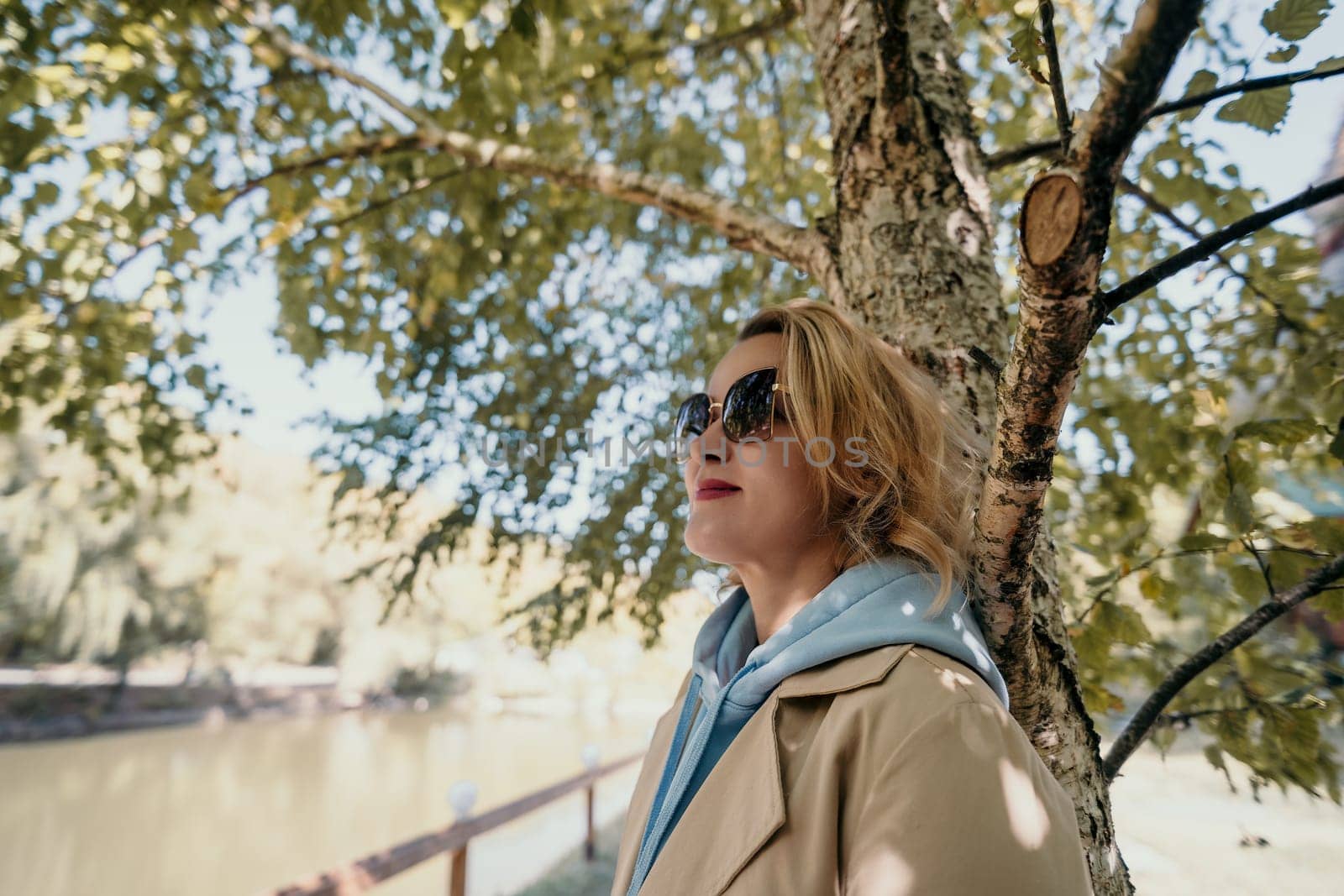 A blonde woman in sunglasses leans against a tree with a serene lake in the background.