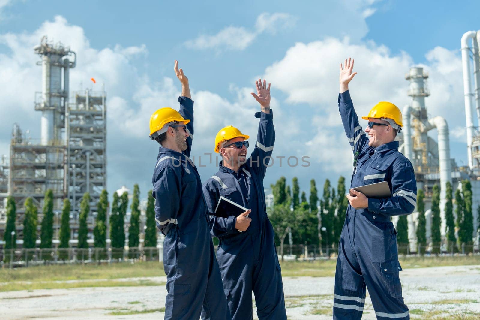 Three factory engineer or technician workers raise hands up to express happiness and stand in front of refining petrochemical factory and tank with day light.