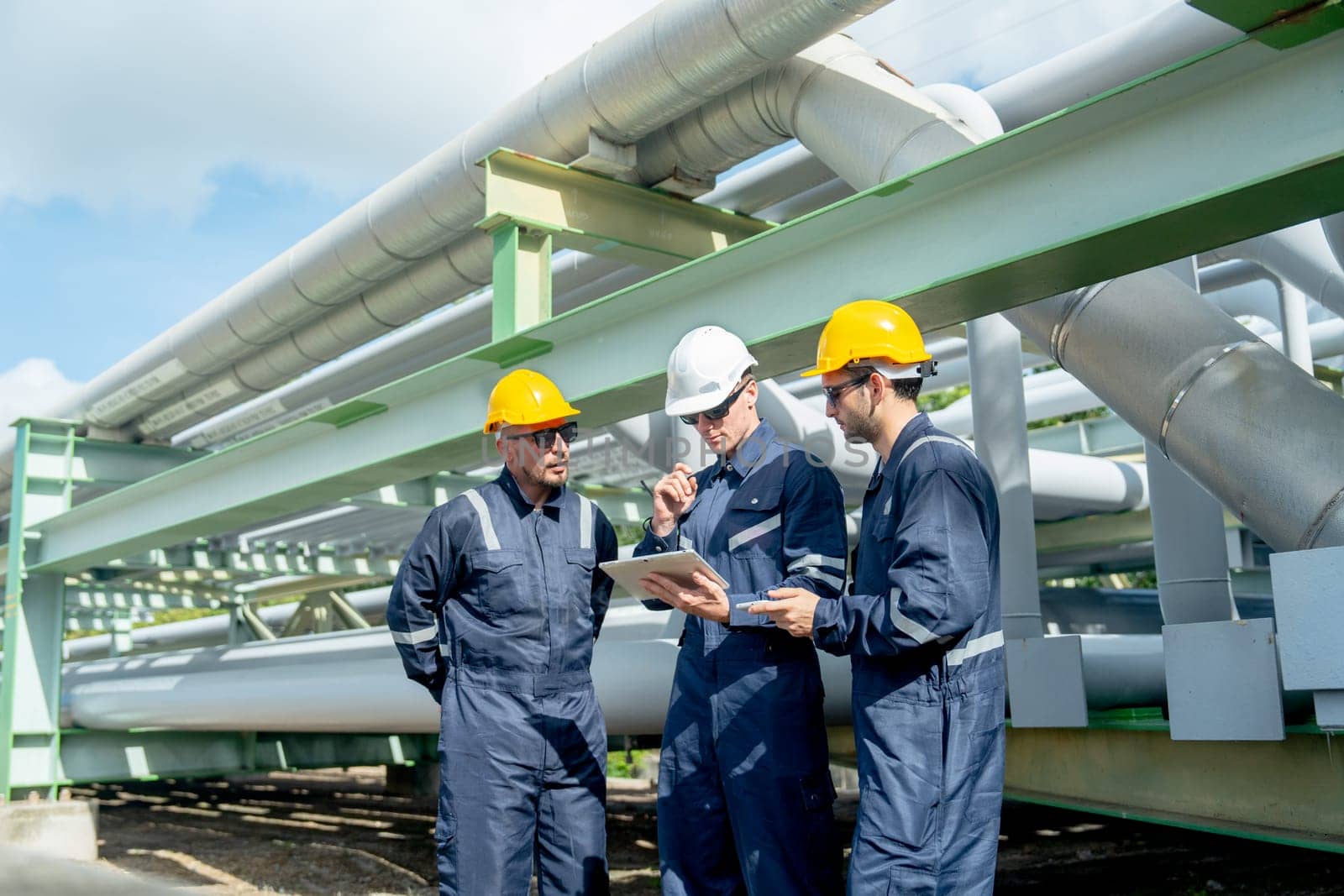 Group of engineer or factory technician worker team stand near petrochemical gas pipeline and use tablet to discuss about their work.