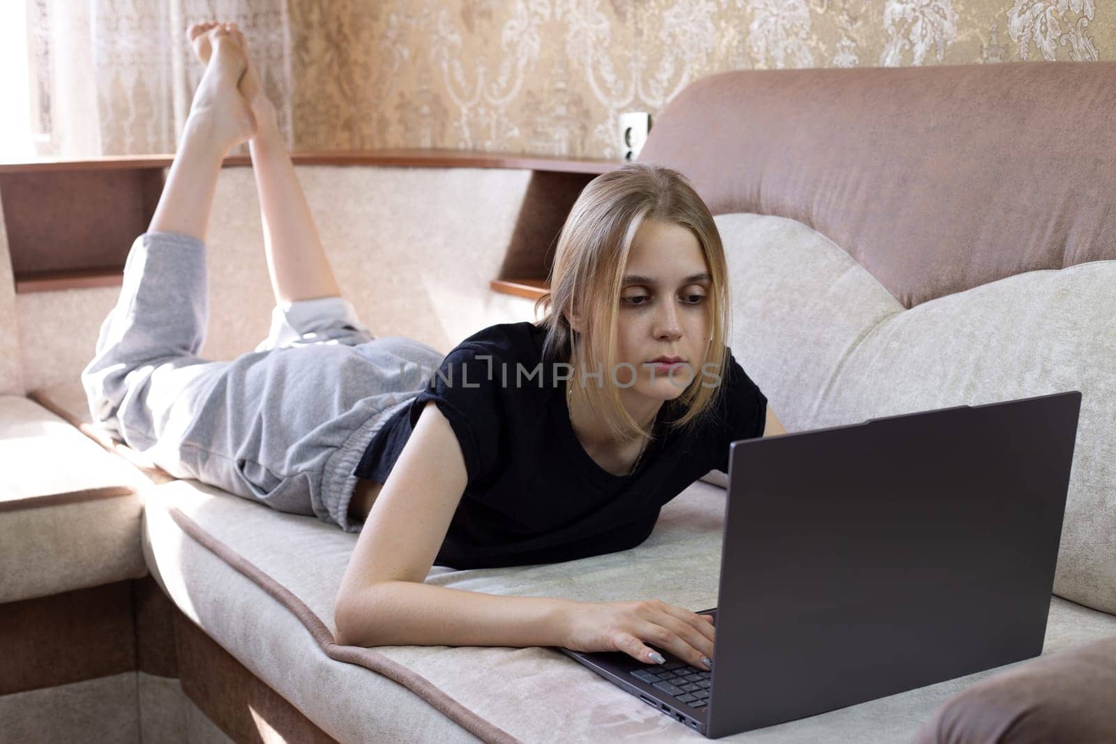 Young girl works at laptop while lying on sofa on her stomach in domestic room. Online office work from home, work of the future from your home room using computer