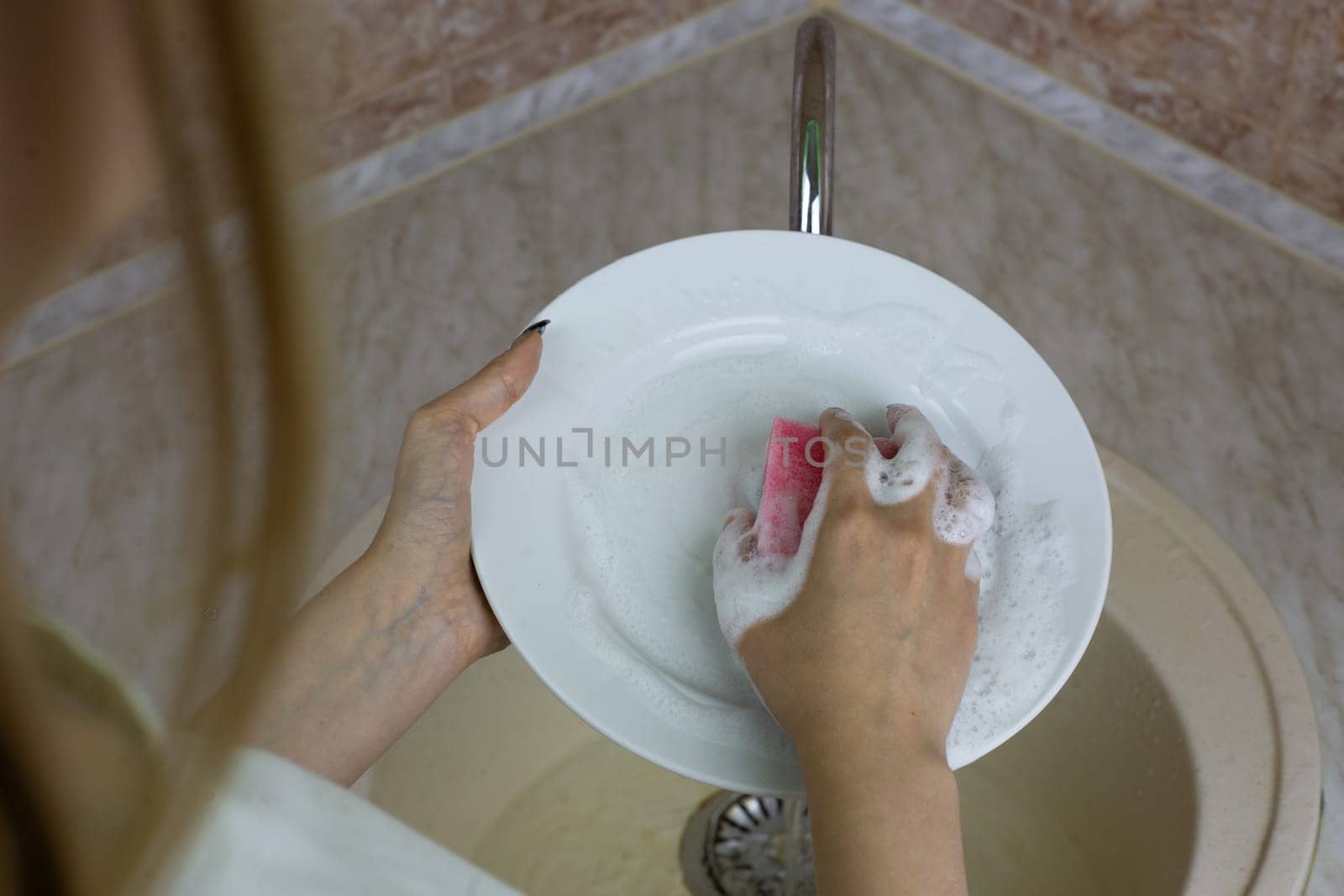 Female hands washing white plate in sink with sponge and foam from detergent in the kitchen at home. Cleaning dishes after meals, daily routine in apartment